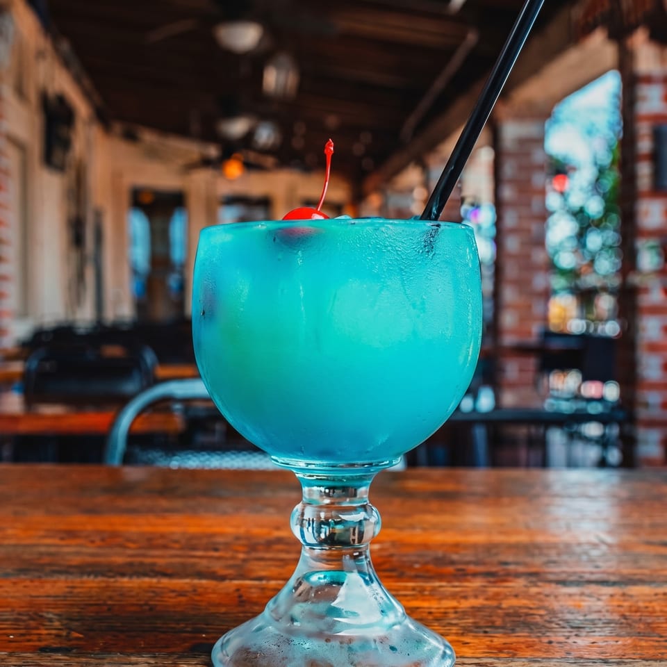 A vibrant blue cocktail with a cherry and straw sits on a wooden table at an outdoor restaurant.