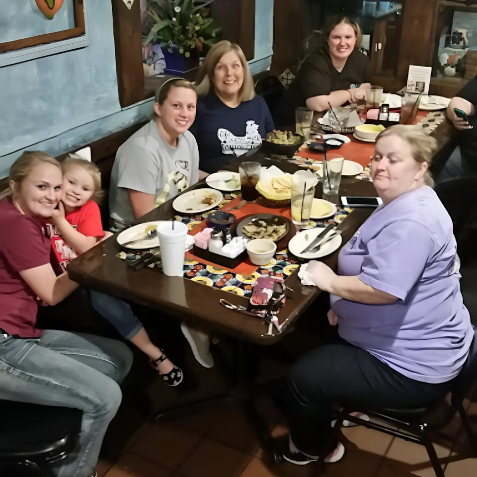 Six women and one child smiling and dining together at a restaurant table with food and drinks.