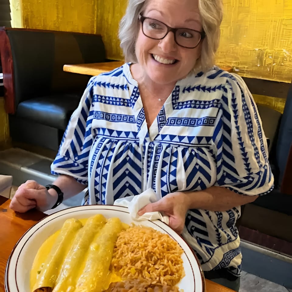 A smiling woman in a blue and white patterned shirt holds a plate of enchiladas and rice.