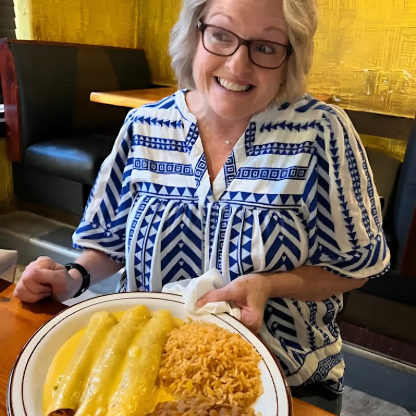 A smiling woman in a blue and white patterned shirt holds a plate of enchiladas and rice.