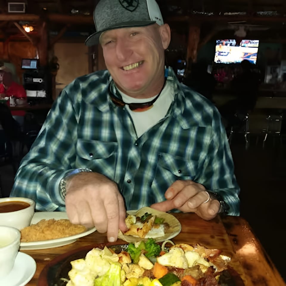 Man in a plaid shirt and hat, smiling while preparing a taco at a restaurant table.
