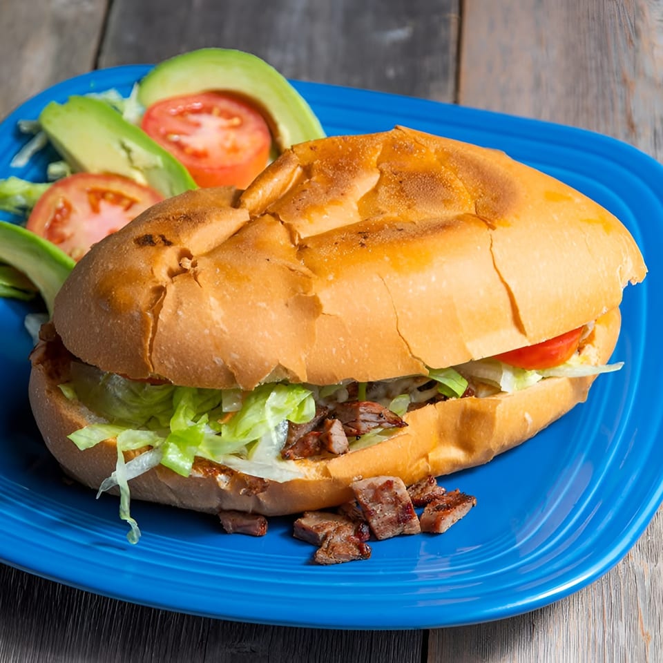 A Mexican torta sandwich with meat, lettuce, and tomato, served with avocado slices on a blue plate.