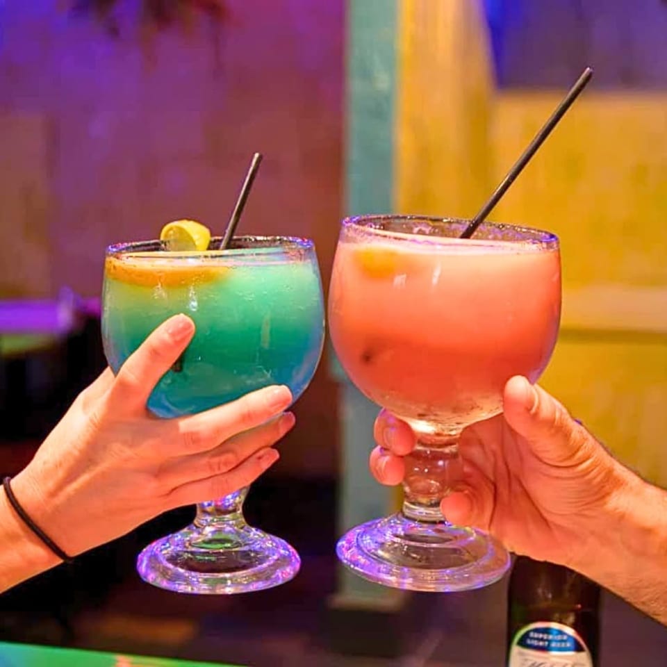 Two hands holding large, colorful margaritas with straws and lemon slices in a vibrant setting.