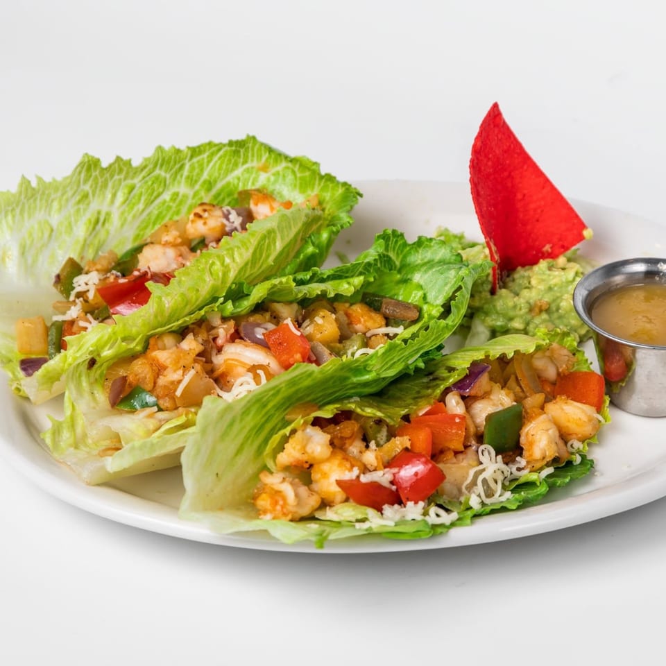 Two lettuce wraps filled with shrimp, peppers, and onions, served with guacamole and sauce.