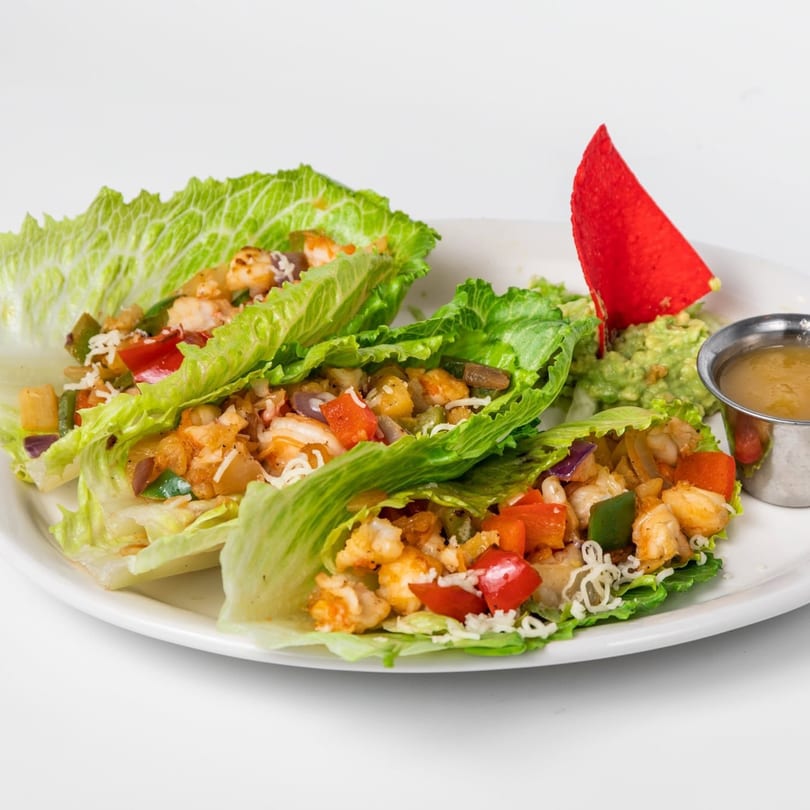 Two lettuce wraps filled with shrimp, peppers, and onions, served with guacamole and sauce.
