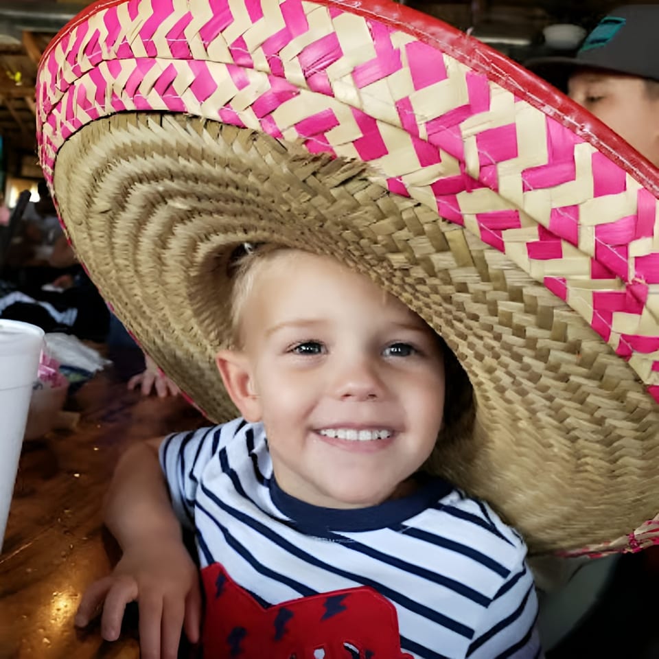 A smiling child wearing an oversized pink and straw sombrero at a restaurant.