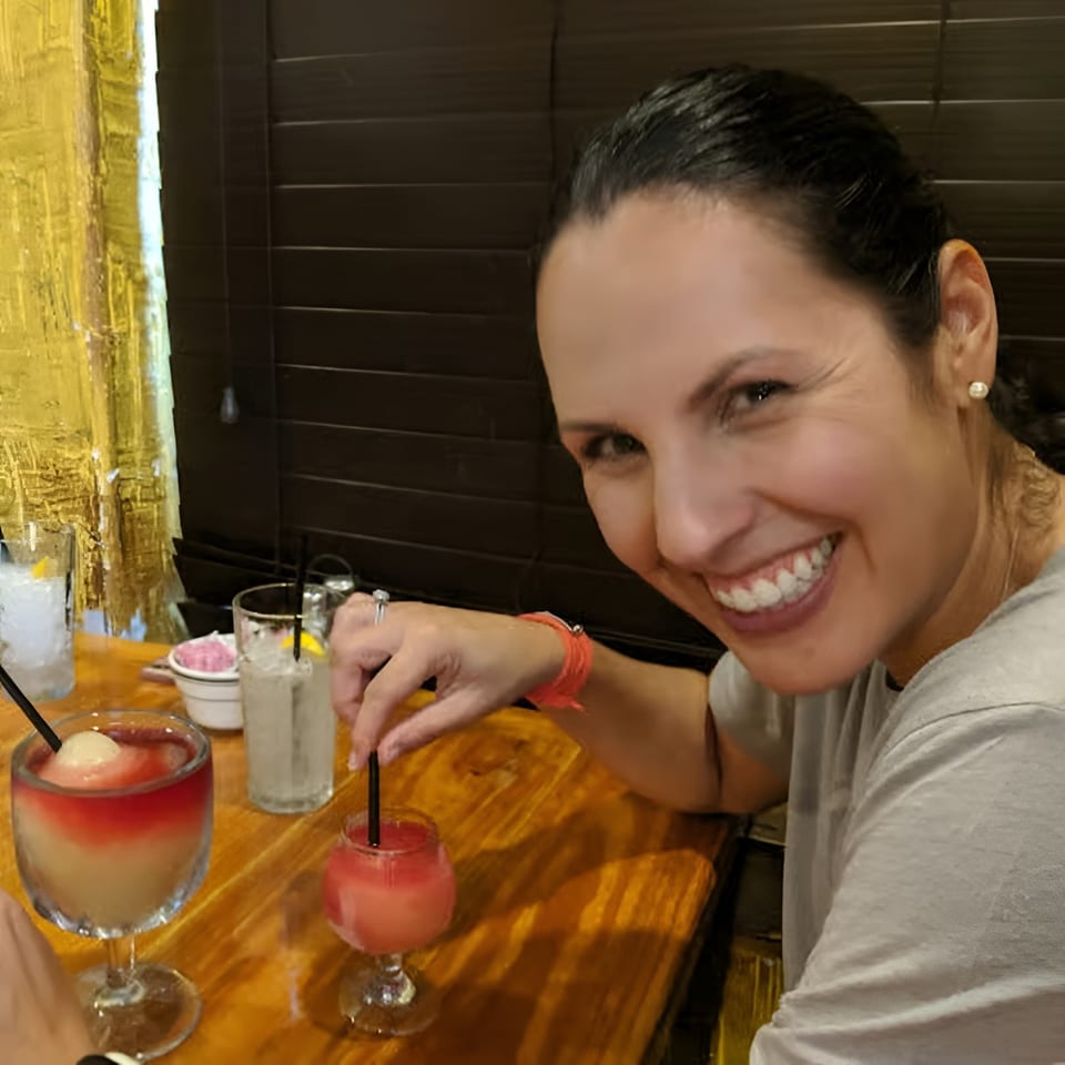 Smiling woman at a restaurant table with several colorful frozen drinks, stirring one with a straw.