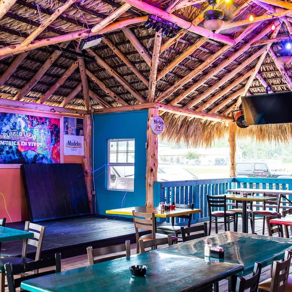 An outdoor restaurant patio with a thatched roof, blue walls, a stage, and empty tables.