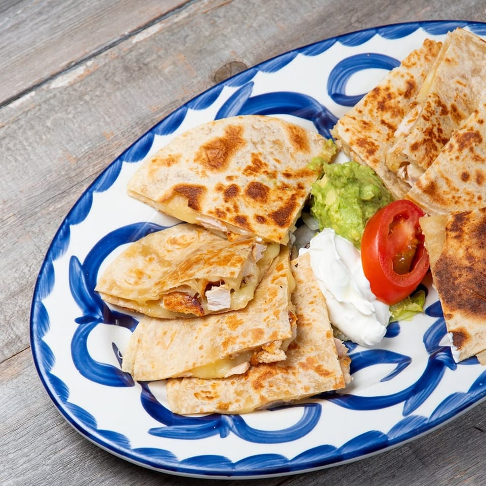 Chicken quesadillas with guacamole, sour cream, and a tomato slice on a blue and white plate.