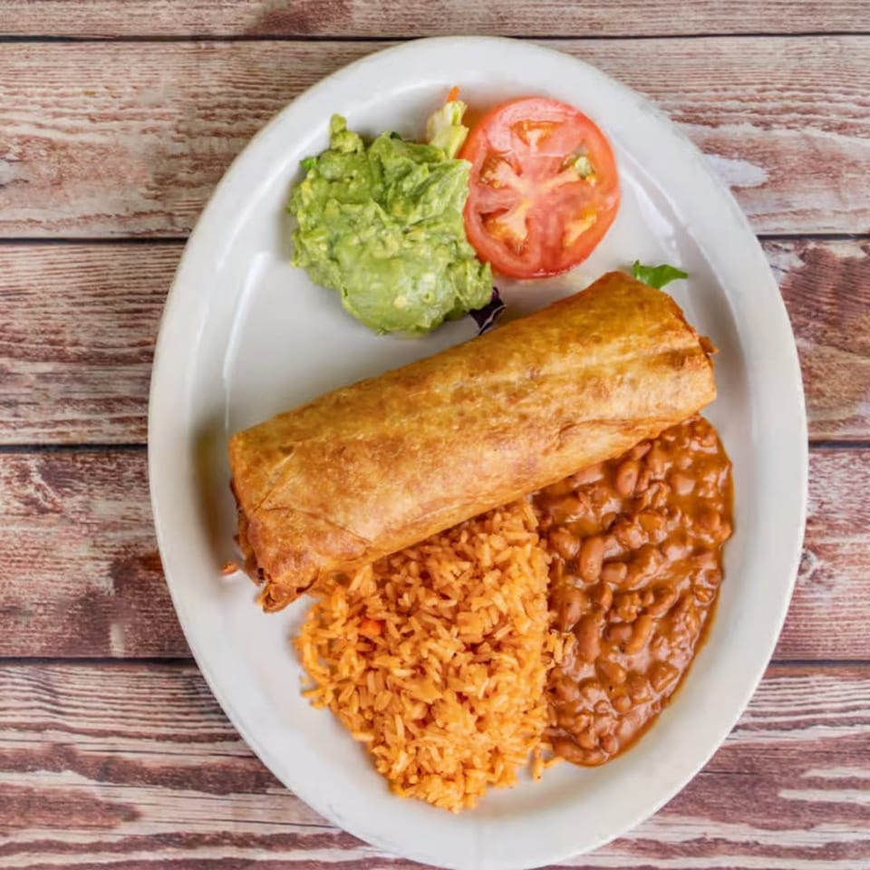 A golden-brown chimichanga, rice, refried beans, guacamole, and tomato slices on a white plate.