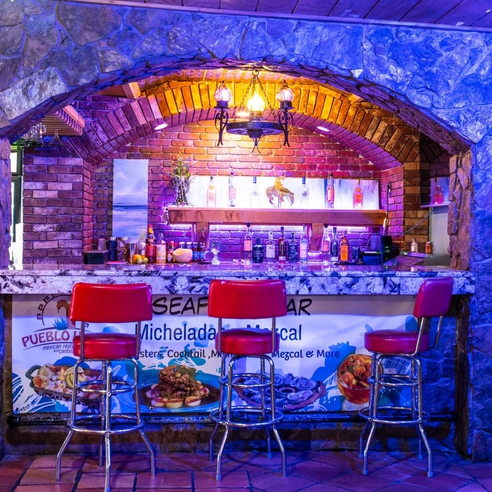 A vibrant bar with red stools, a stone and brick arch, and bottles lit by blue and purple lights.