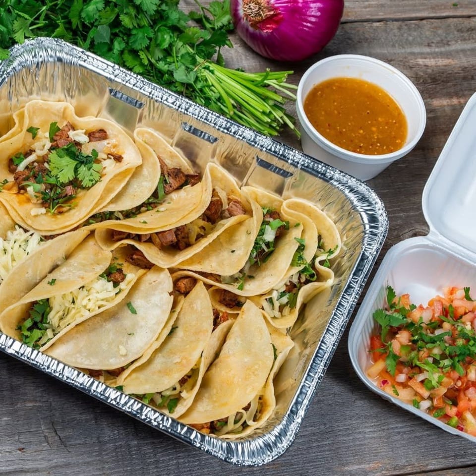 A foil tray of tacos with meat, cilantro, and onions, alongside salsa and pico de gallo.