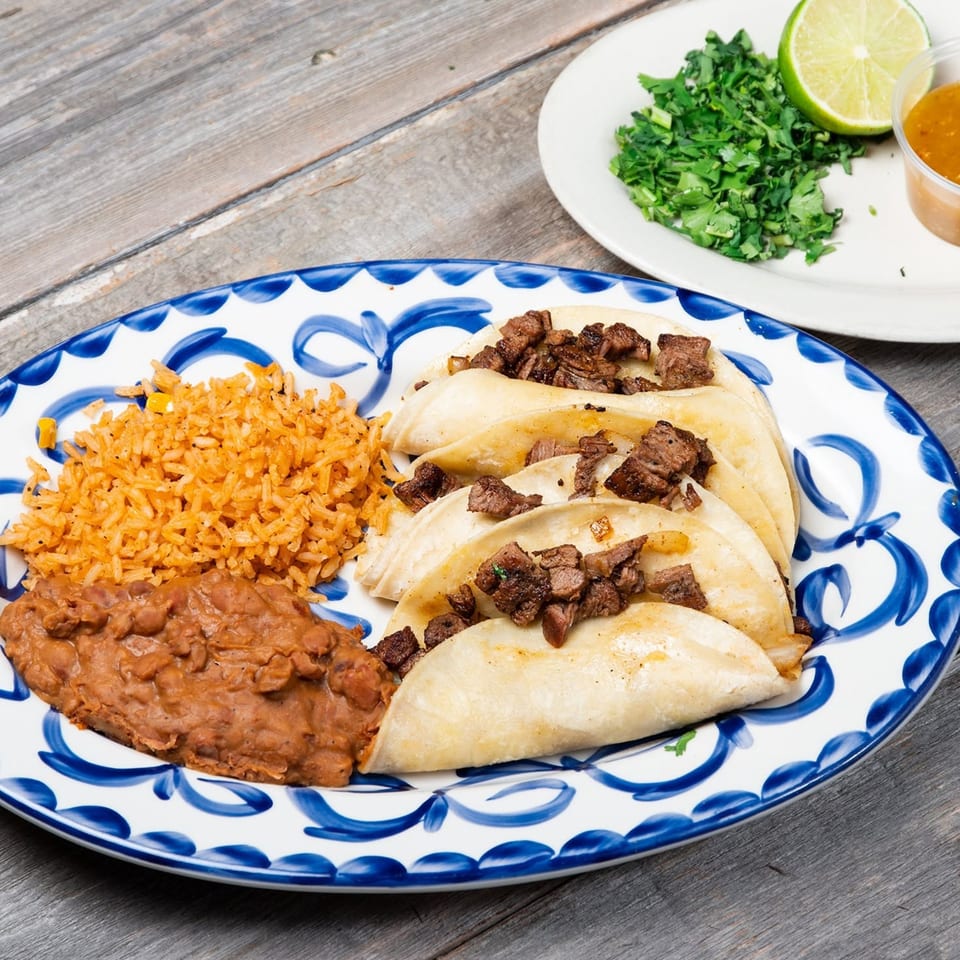 Four beef tacos with rice and refried beans on a blue and white plate, with cilantro and lime.