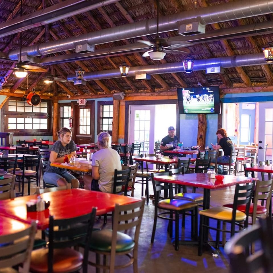 Restaurant interior with patrons dining at red tables, exposed ceiling ducts, and a TV showing sports.