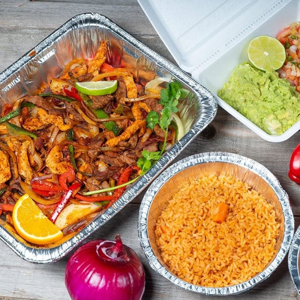 Fajitas with chicken, steak, peppers, and onions in a foil pan, with rice, guacamole, and salsa.