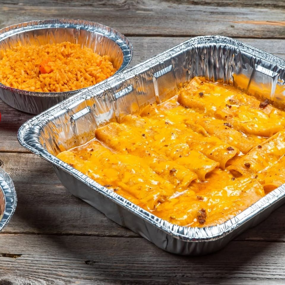 A large aluminum tray of cheesy enchiladas next to a smaller tray of orange rice on a wooden table.